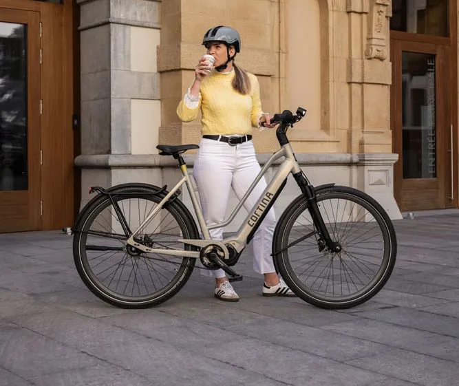 woman with helmet standing next to a cortina e-silento women's bike in purse grey.